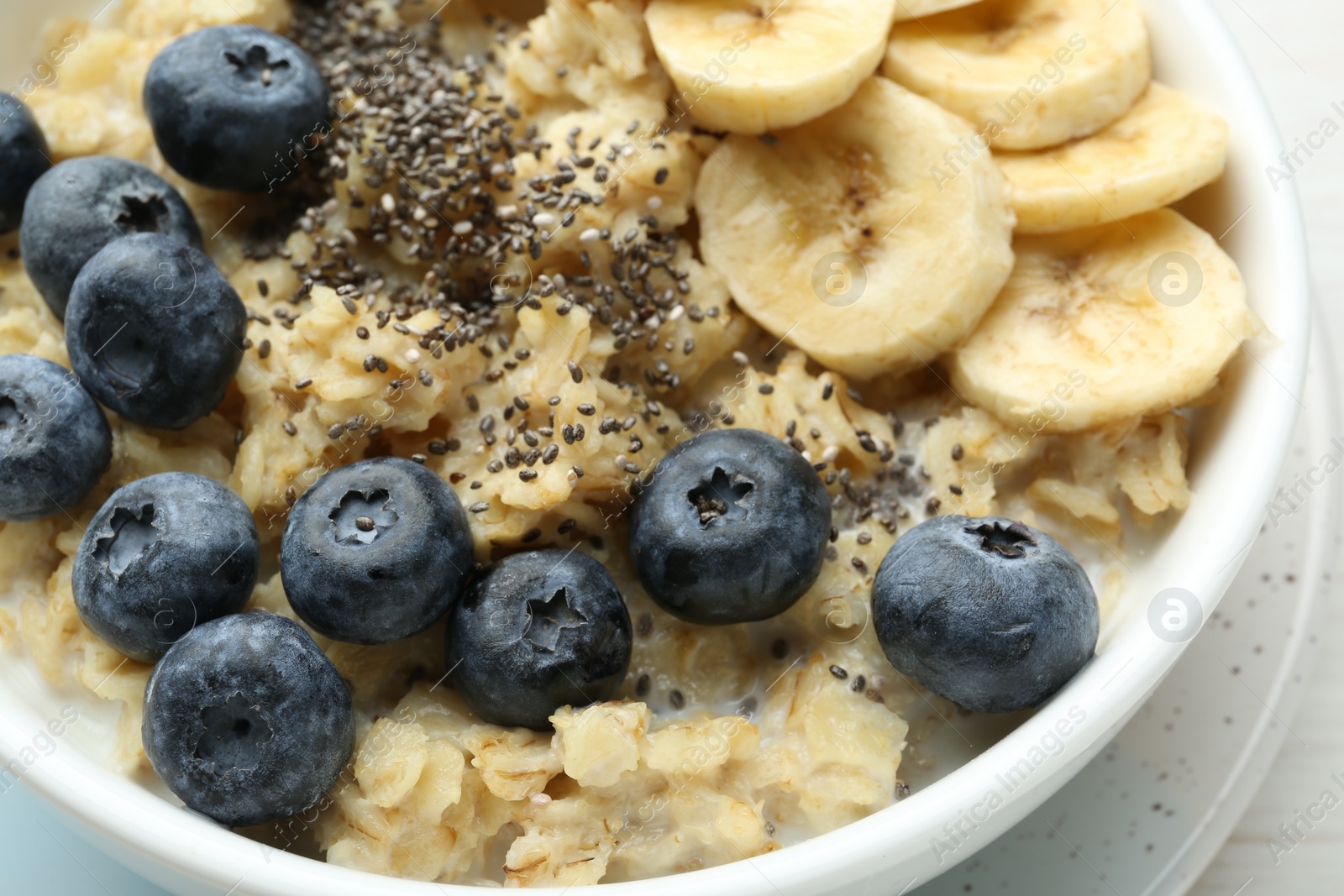 Photo of Tasty oatmeal with banana, blueberries and chia seeds served in bowl on white table, closeup