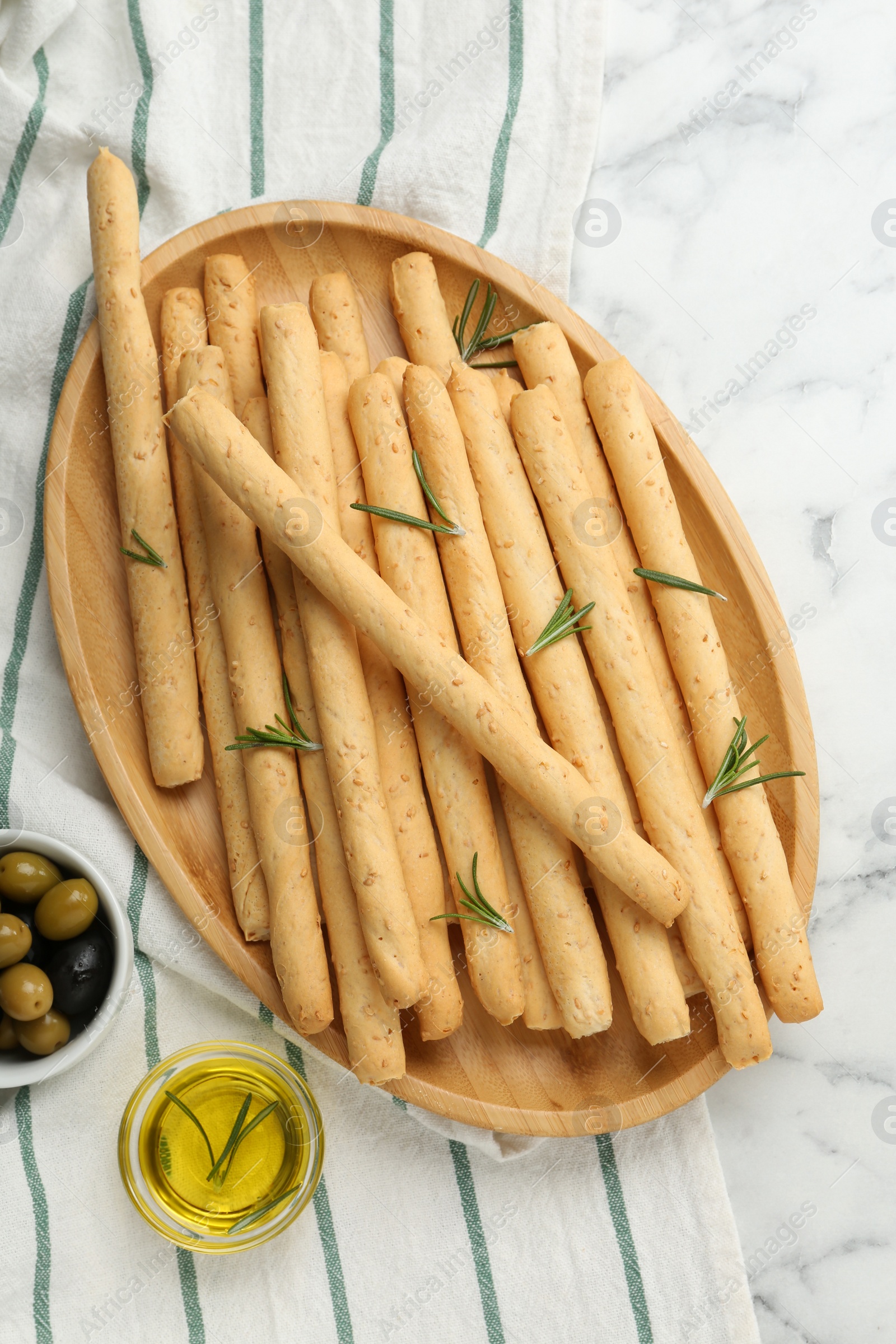 Photo of Delicious grissini, rosemary, oil and olives on white marble table, flat lay