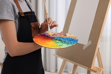 Photo of Woman mixing paints on palette with brush near easel in studio, closeup