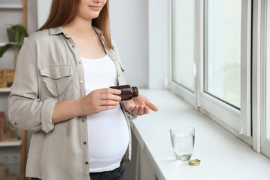Pregnant woman taking pill from bottle near window at home, closeup