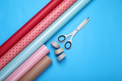 Photo of Rolls of wrapping papers, scissors and ribbons on light blue background, flat lay. Space for text
