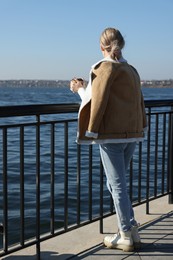 Photo of Lonely woman with cup of drink on pier near river, back view