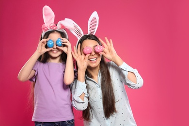 Mother and daughter in bunny ears headbands holding Easter eggs near eyes on color background