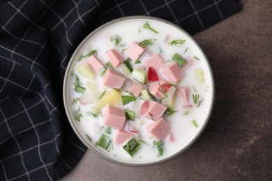 Delicious cold summer soup (okroshka) with boiled sausage in bowl on brown table, top view