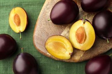 Many tasty ripe plums and wooden board on green fabric, flat lay