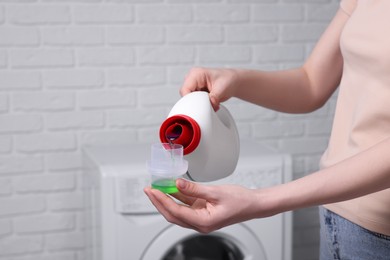 Photo of Woman pouring fabric softener from bottle into cap for washing clothes near white brick wall, closeup