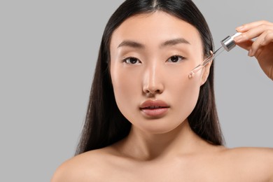 Beautiful young woman applying cosmetic serum onto her face on grey background, closeup. Space for text