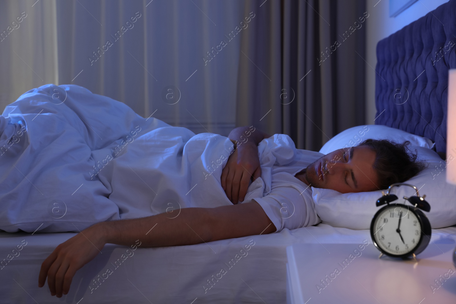 Photo of Alarm clock on nightstand near sleeping young man. Bedtime