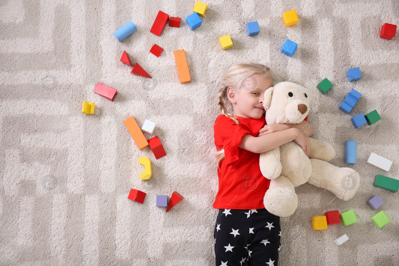 Photo of Cute little girl lying on cozy carpet with cubes and teddy bear at home