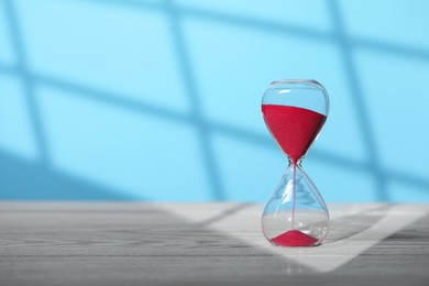 Photo of Hourglass with red flowing sand on white wooden table against light blue background, space for text