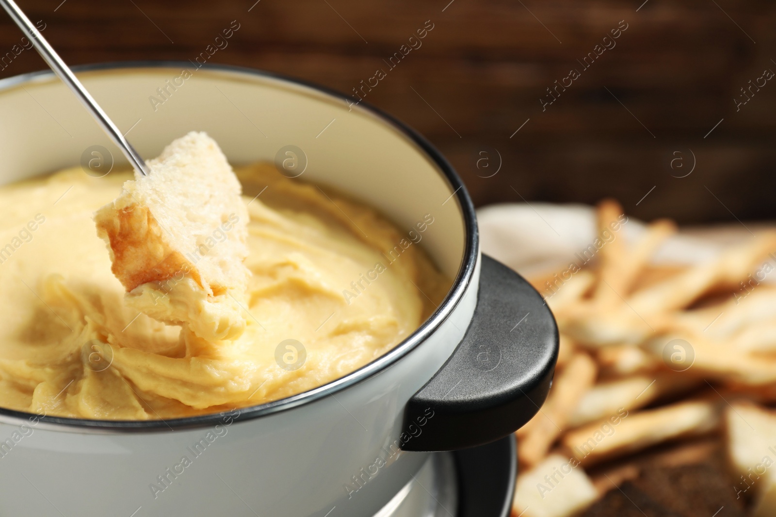 Photo of Dipping piece of bread into fondue pot with melted cheese on blurred background, closeup