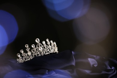 Photo of Beautiful silver tiara with diamonds on dark cloth against blurred lights, space for text