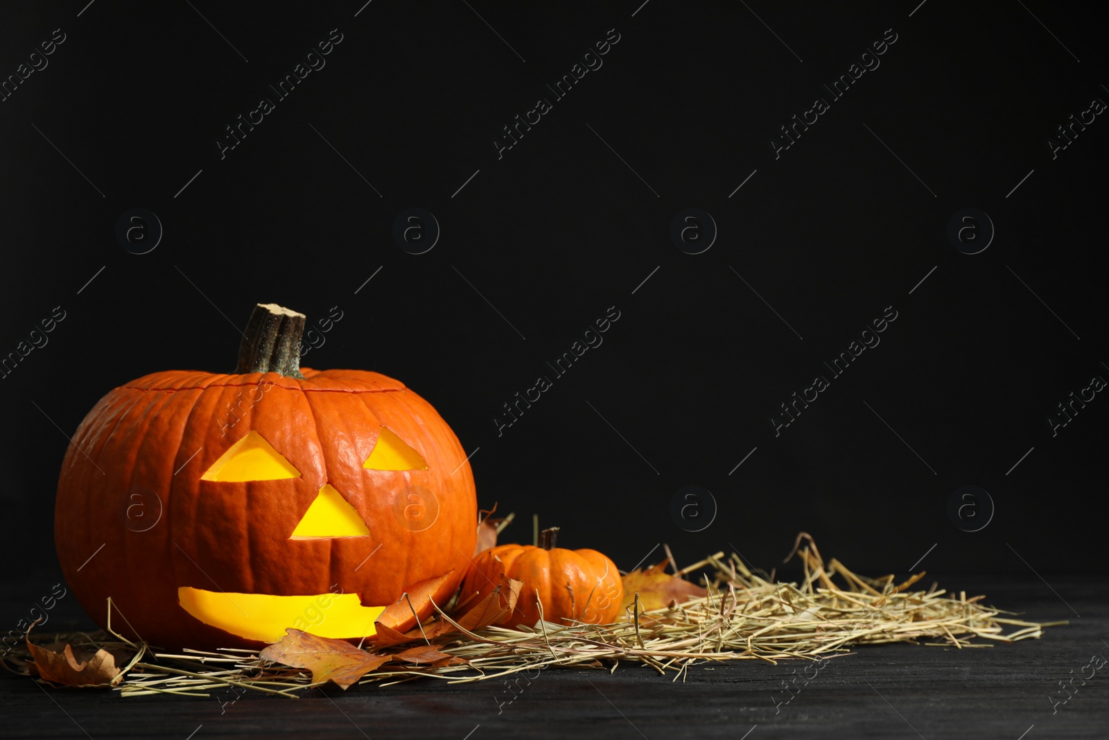 Photo of Pumpkin jack o'lantern, straw and autumn leaves on table in darkness, space for text. Halloween decor