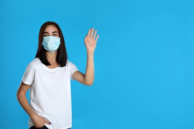 Young woman in protective mask showing hello gesture on light blue background, space for text. Keeping social distance during coronavirus pandemic