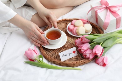 Photo of Tasty breakfast served in bed. Woman with tea, macarons, gift box, flowers and I Love You card at home, closeup