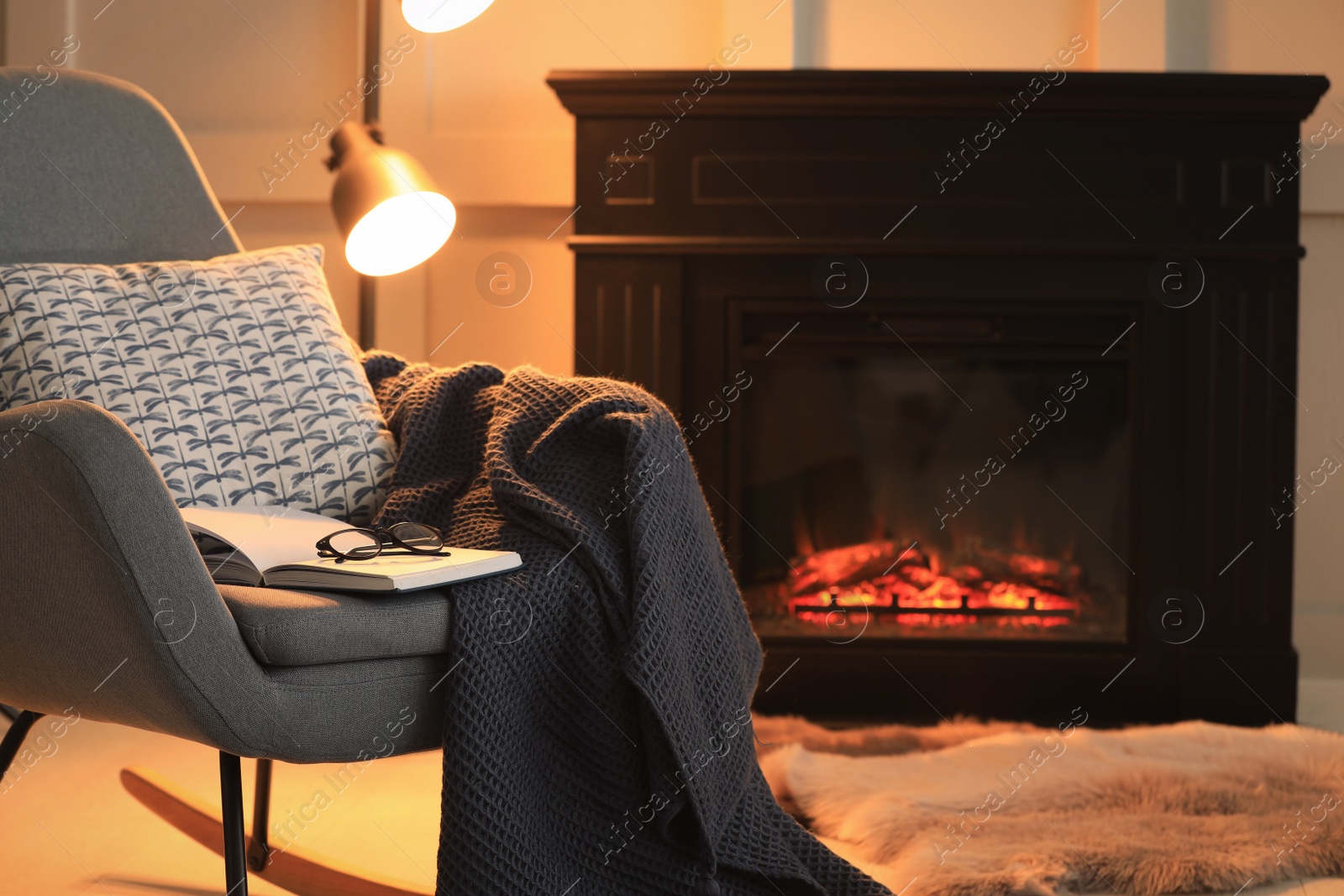 Photo of Open book, glasses and plaid on rocking chair near fireplace at home, space for text. Cozy atmosphere