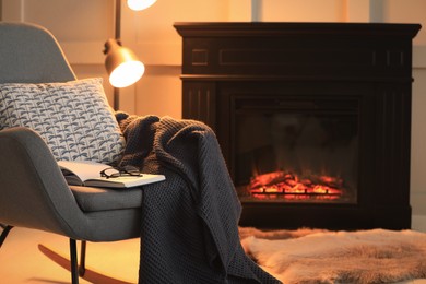 Photo of Open book, glasses and plaid on rocking chair near fireplace at home, space for text. Cozy atmosphere