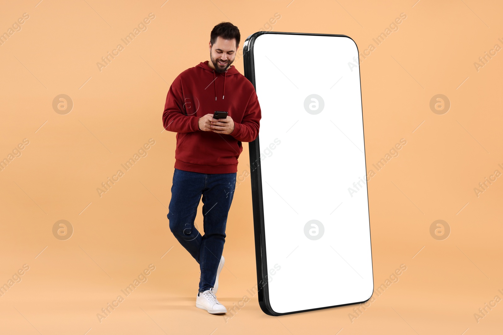 Image of Man with mobile phone standing near huge device with empty screen on dark beige background. Mockup for design