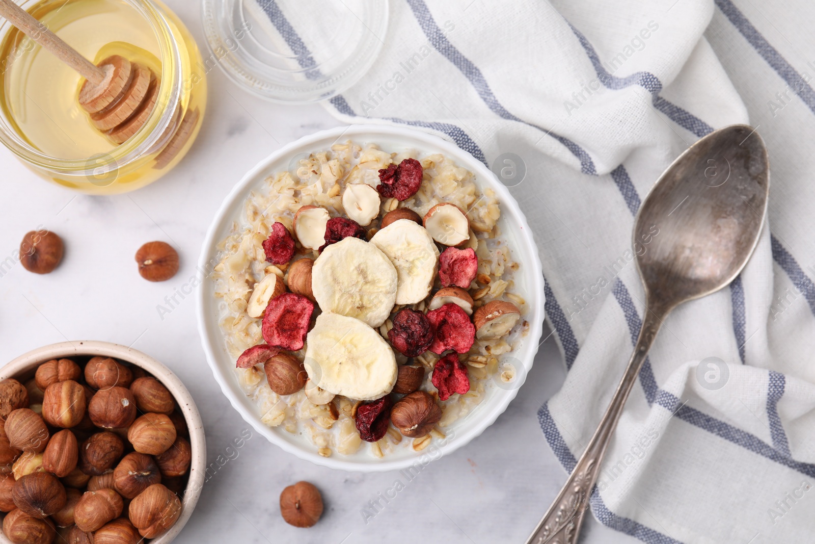 Photo of Delicious oatmeal with freeze dried berries, banana and hazelnuts served on white marble table, flat lay