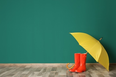 Photo of Colorful umbrella and rubber boots on floor against green wall. Space for text