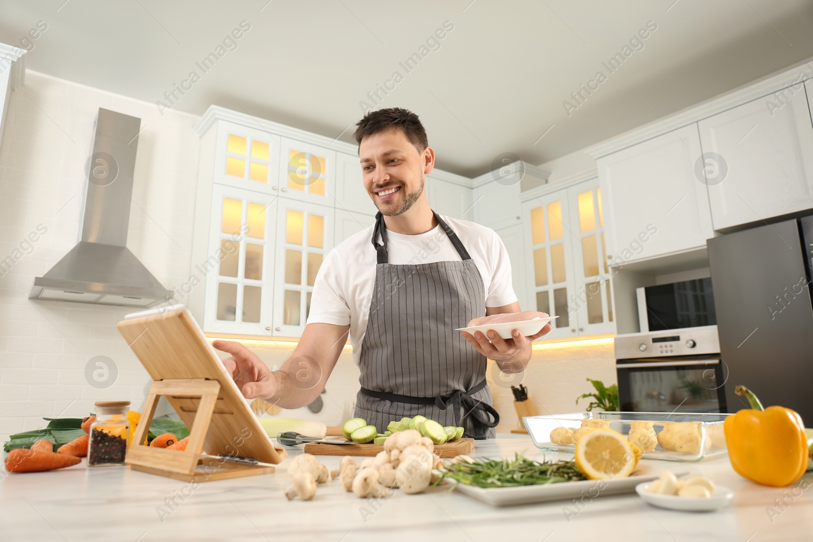 Photo of Happy man making dinner while watching online cooking course via tablet in kitchen