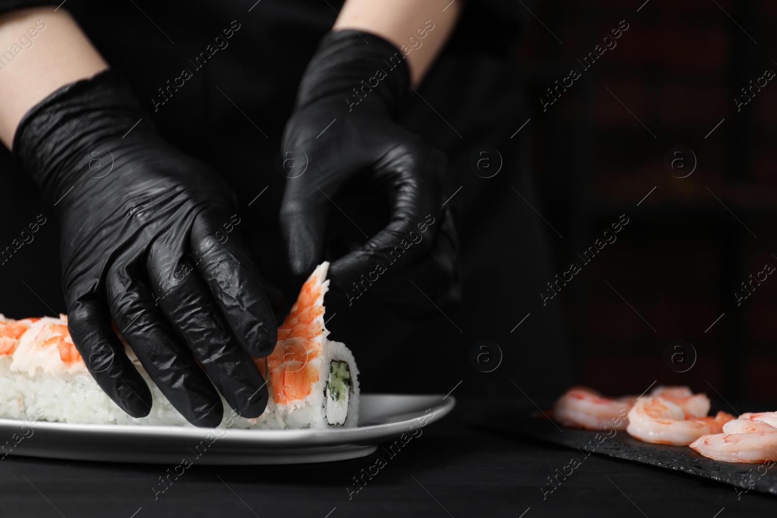 Photo of Chef in gloves making sushi rolls with shrimps at black wooden table, closeup