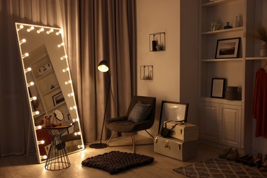 Makeup room. Stylish mirror with light bulbs, beauty products on table and armchair indoors