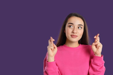 Photo of Woman with crossed fingers on purple background, space for text. Superstition concept
