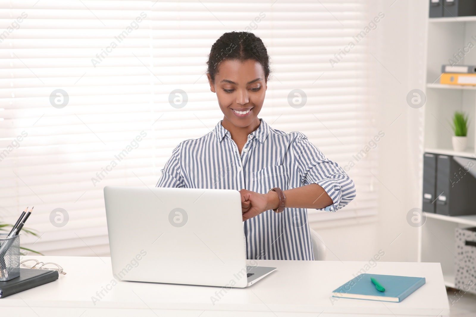 Photo of Smiling African American intern checking time on watch while working at white table in office