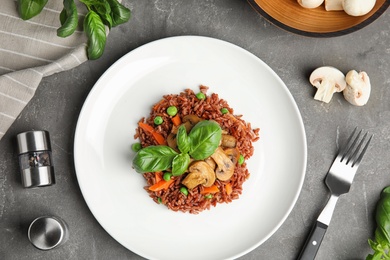 Photo of Plate of tasty brown rice with vegetables and mushrooms on grey table, flat lay