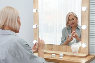 Portrait of charming mature woman with healthy beautiful face skin and natural makeup applying cream indoors