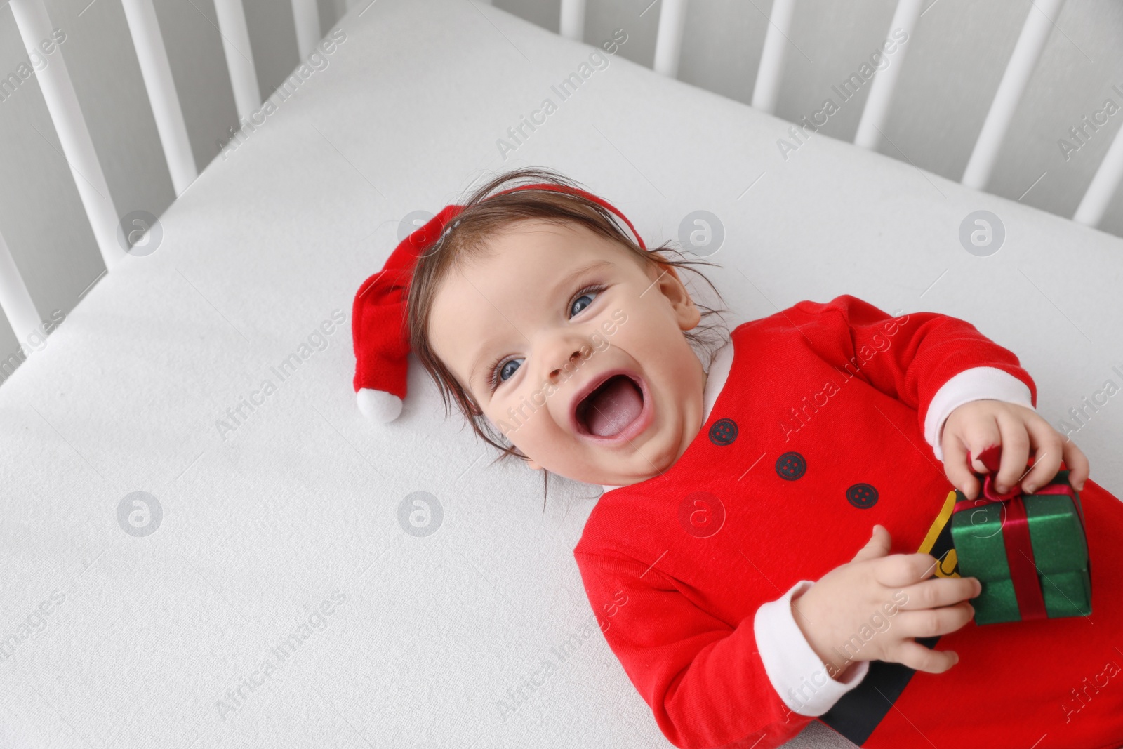 Photo of Cute baby wearing festive Christmas costume with gift box in crib, above view