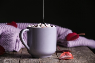 Pouring chocolate topping onto hot cozy drink in cup near autumn leaves on wooden table. Space for text