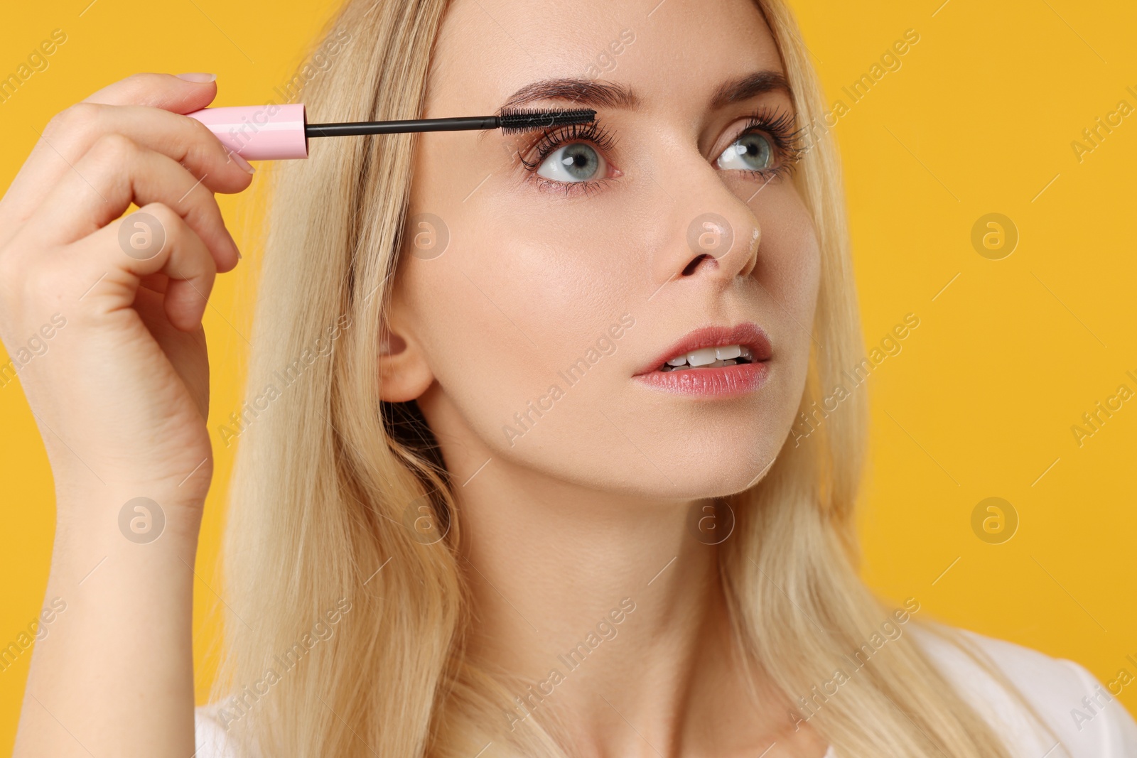 Photo of Beautiful woman applying mascara on orange background, closeup