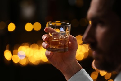 Man holding glass of whiskey with ice cubes against blurred lights, selective focus