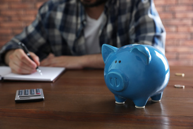 Photo of Man counting money at wooden table, focus on piggy bank