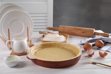 Pie tin with fresh dough, rolling pin and ingredients on white wooden table. Making quiche