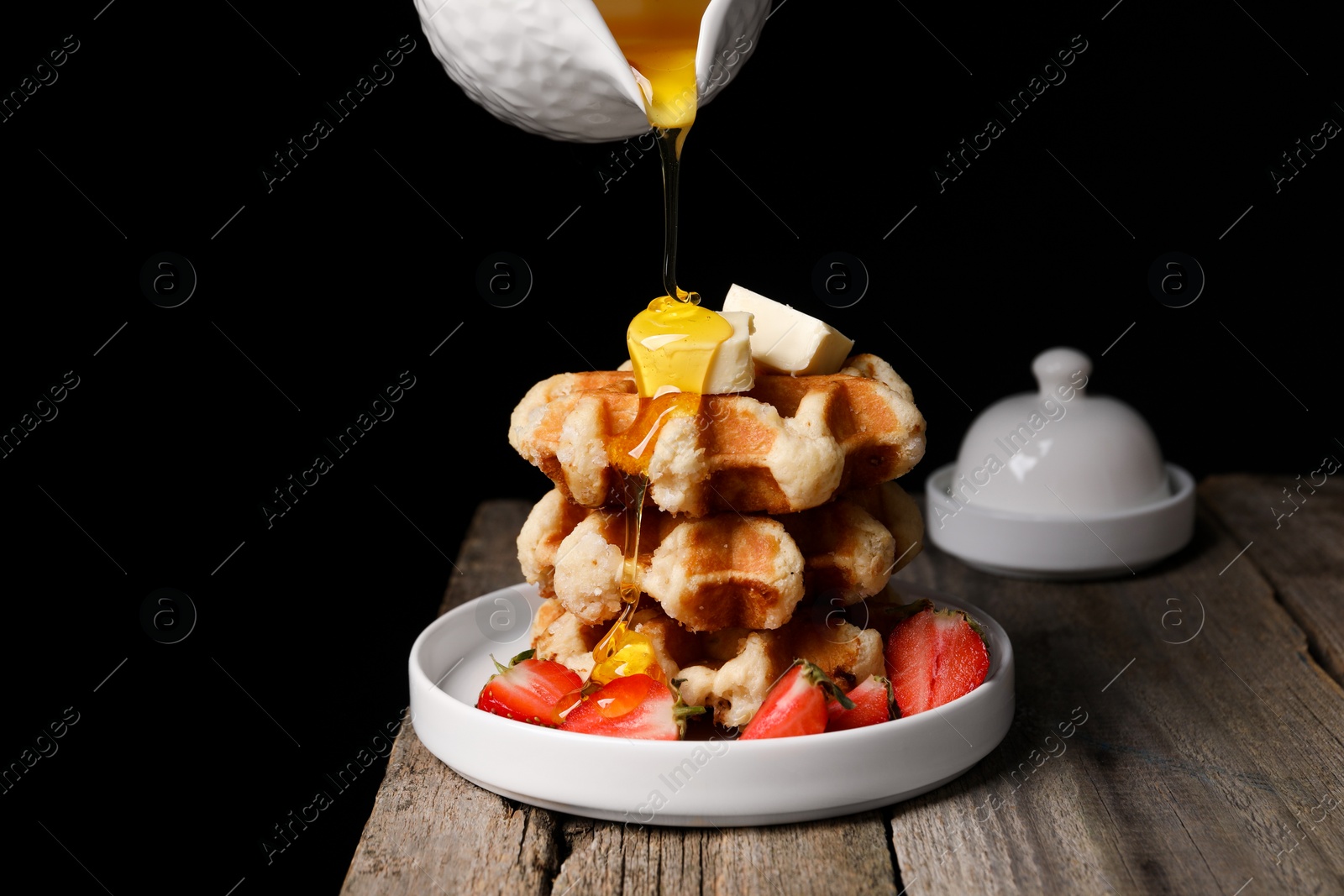 Photo of Pouring honey onto delicious Belgian waffles with strawberries and butter at wooden table against black background, space for text