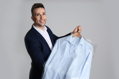 Photo of Man holding hanger with shirt in plastic bag on light grey background. Dry-cleaning service