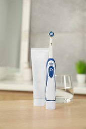 Electric toothbrush, tube with paste and glass of water on wooden table in bathroom