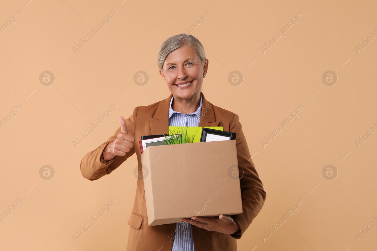 Photo of Happy unemployed senior woman with box of personal office belongings showing thumb up on beige background