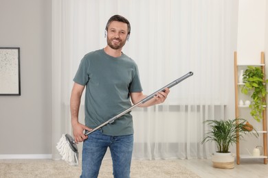 Photo of Enjoying cleaning. Happy man in headphones with mop at home, space for text