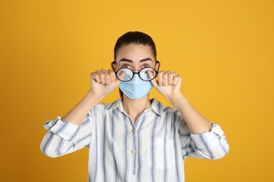 Woman wiping foggy glasses caused by wearing medical mask on yellow background
