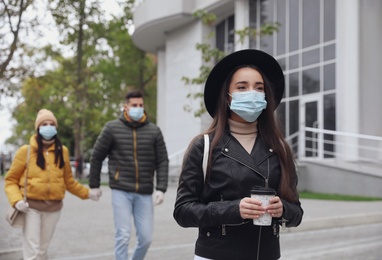 Photo of Young woman in medical face mask with cup of coffee walking outdoors. Personal protection during COVID-19 pandemic