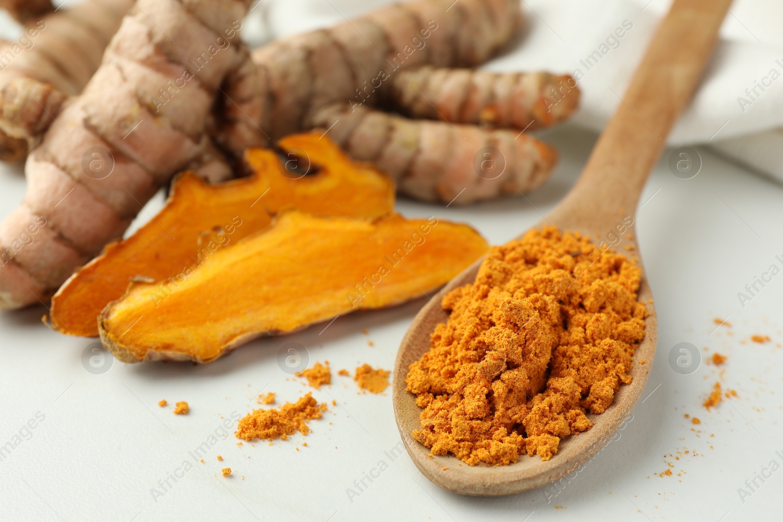 Photo of Wooden spoon with aromatic turmeric powder and raw roots on white table, closeup