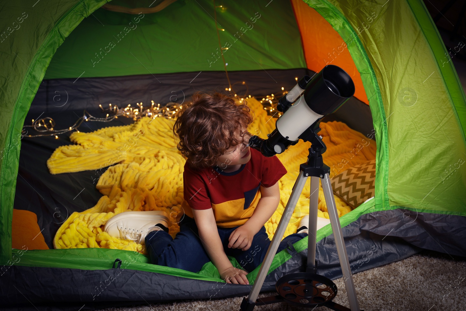 Photo of Little boy looking at stars through telescope while sitting in camping tent indoors