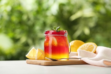 Board with jar of colorful jelly, spoon and lemon slices on white table