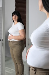 Overweight woman in tight t-shirt and trousers near mirror at home