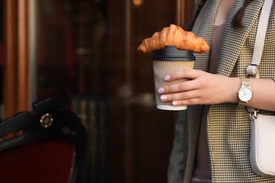 Woman holding tasty croissant and cup of coffee near wooden door outdoors, closeup