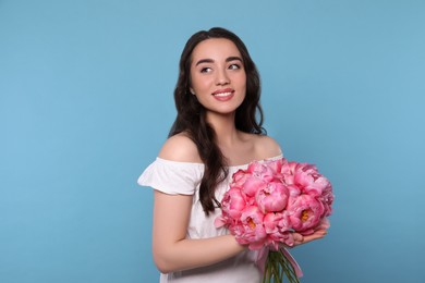 Beautiful young woman with bouquet of pink peonies on light blue background